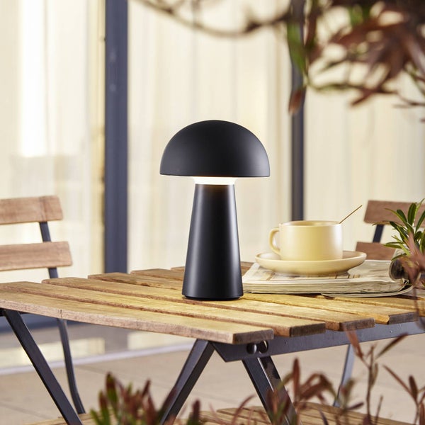 LED-Tischleuchte in Pilzform auf einem hölzernen Gartentisch auf einer hellen Terrasse mit Tasse und Zeitung.