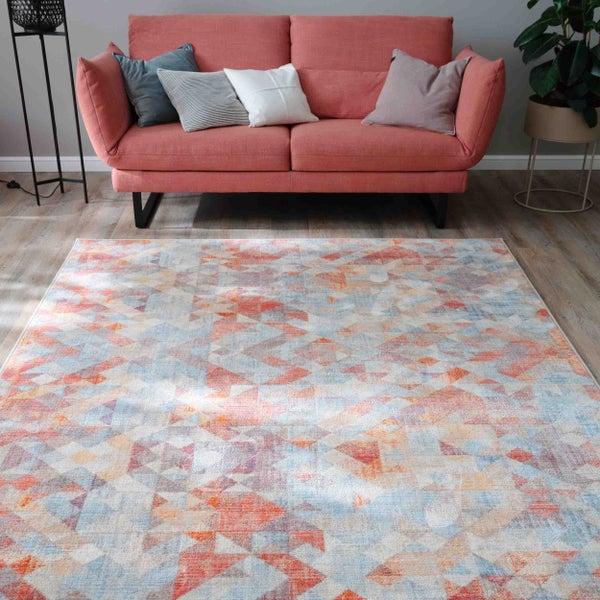 Moderner Teppich mit geometrischem Dreiecksmuster vor einem korallenfarbenen Sofa auf Holzboden in einem hellen Wohnzimmer.
