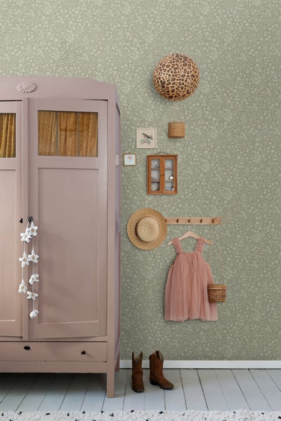 Hell eingerichtetes Zimmer im Vintage-Stil mit floraler Vliestapete, einem rosa Kleiderschrank aus Holz und einer Hakenleiste mit Kinderkleid.