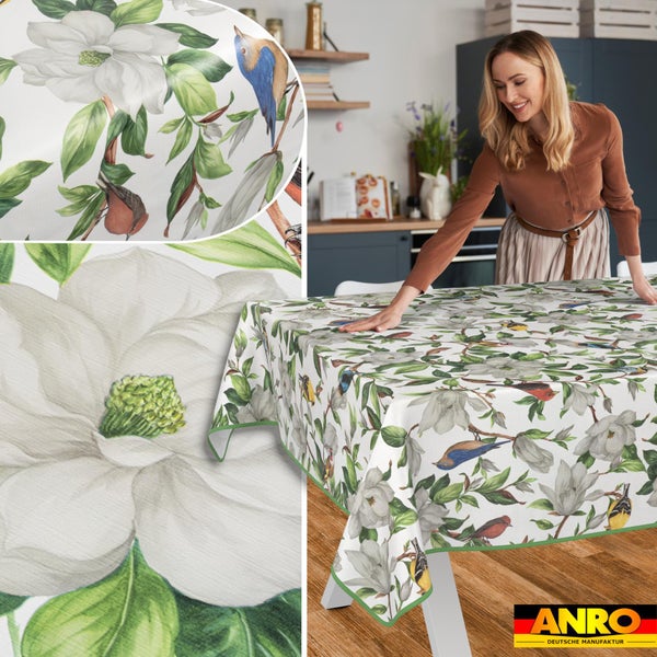 Tischdecke mit Magnolien und Vögeln, die auf einem Esstisch in einer hellen, organisierten Küche ausgebreitet wird. ANRO Logo.