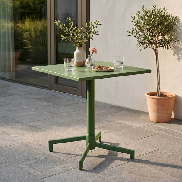 Moderner, quadratischer Gartentisch aus grünem Metall auf einer Steinterrasse, dekoriert mit Gläsern und Vasen, neben einem Olivenbaum im Topf.