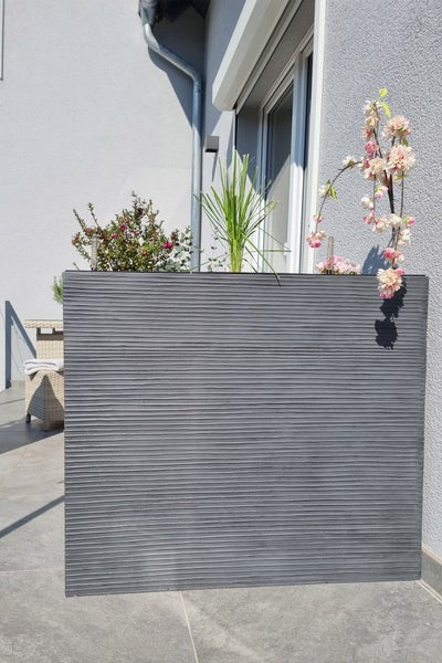 Hoher rechteckiger Pflanzkübel in Anthrazit mit horizontaler Rillenstruktur auf einer Terrasse, bepflanzt mit verschiedenen Gartenpflanzen.