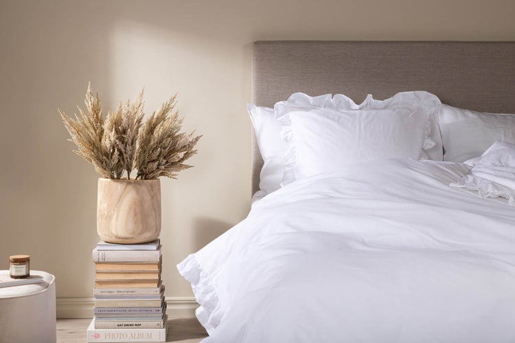Schlafzimmer mit weißer Rüschenbettwäsche, grauem Polsterkopfteil und einem Stapel Bücher als Beistelltisch mit Trockengras in einer Holzvase.
