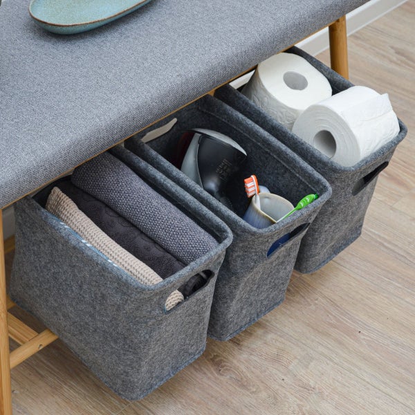 Drei graue Filzkörbe mit Handtüchern, Haartrockner und Toilettenpapier, ordentlich verstaut unter einer gepolsterten Holzbank.