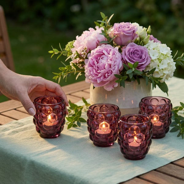 Hand platziert ein strukturiertes Windlicht aus Glas mit brennender Kerze auf einem Gartentisch neben weiteren Windlichtern und einem Blumenstrauß.