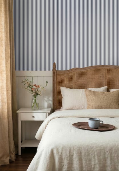 Schlafzimmer mit blau-weiß gestreifter Tapete, Holzbett mit Kopfteil aus Geflecht und weißem Nachttisch mit Blumenvase.