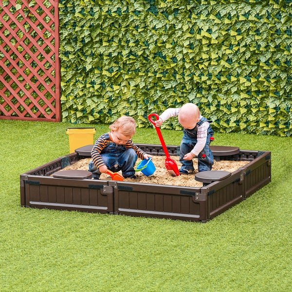 Zwei Kinder spielen in einem braunen Sandkasten mit Abdeckung im Garten.