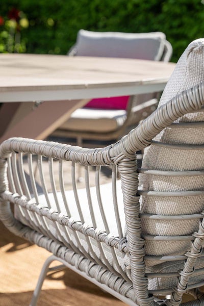 Nahaufnahme eines modernen Gartenstuhls aus grauem Kunststoffgeflecht mit weichem Sitzkissen an einem hellen Holztisch im Garten.