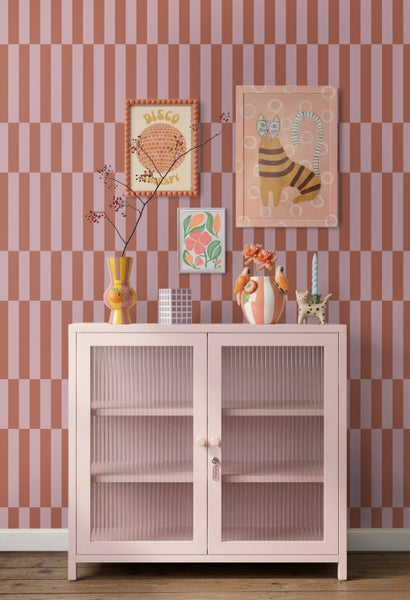 Rosa Metallschrank mit geriffelten Glastüren vor gemusterter Tapete mit dekorativen Vasen und Bildern in hellem, modernem Ambiente.