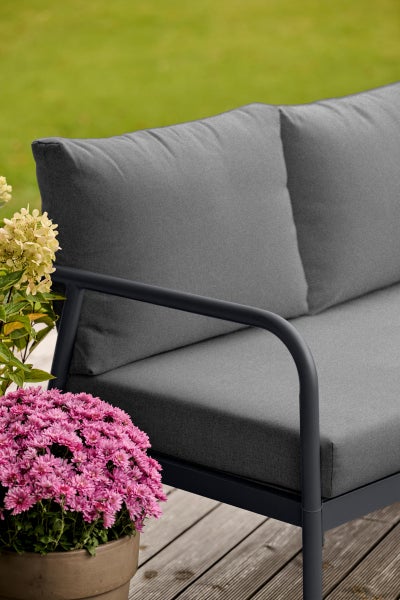 Detailansicht eines grauen Gartensofas mit Metallrahmen und Polstern neben rosa Blumen auf einer Holzterrasse.
