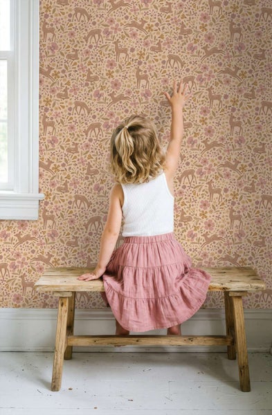 Kind sitzt auf einer Holzbank vor einer Kindertapete mit einem Waldmuster aus Rehen und Blumen in einem hellen Kinderzimmer.