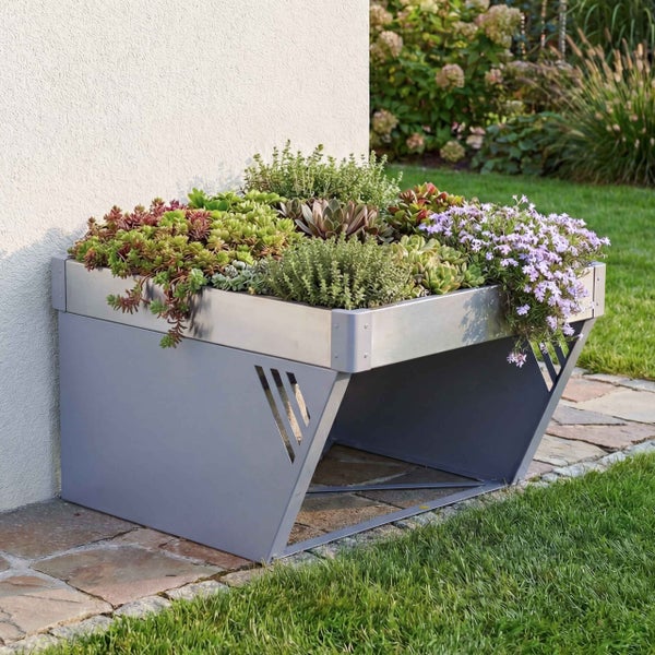 Modernes Hochbeet aus Metall mit Sukkulenten und Blumen bepflanzt, steht auf einem Steinweg an einer Hauswand im Garten.