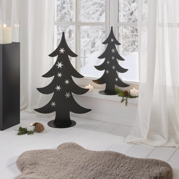Zwei dekorative Metall Tannenbäume mit Sternenaussparungen als weihnachtliche Dekoration in einem hellen Wohnraum vor einem Fenster.
