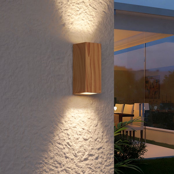 Rechteckige Außenwandleuchte in Holzoptik mit Licht nach oben und unten an einer strukturierten Fassade neben einem modernen Innenraum mit Glasfront.