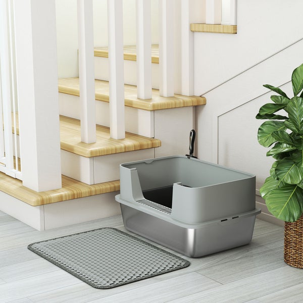 Offene Katzentoilette aus Edelstahl mit hohem Rand, Schaufel und Vorlegematte neben einer Holztreppe in einem hellen Raum.