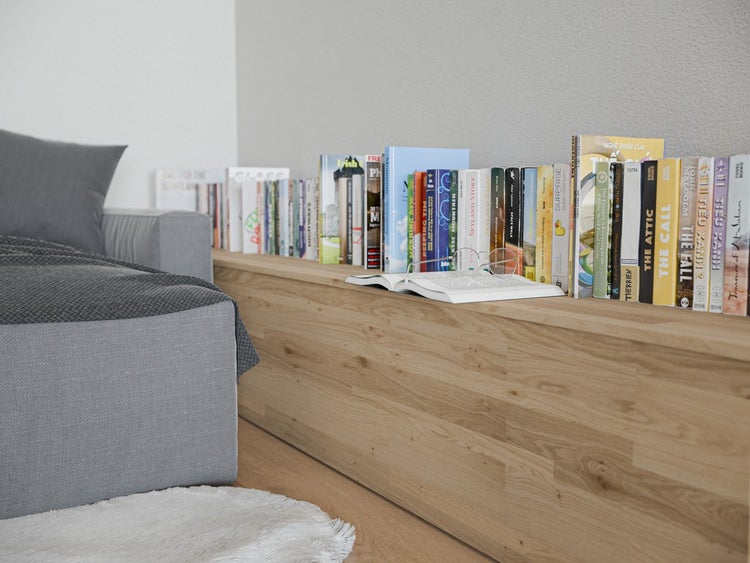 Niedriges Holzregal aus heller Eiche mit Büchern neben einem Sofa in einem hellen Raum.