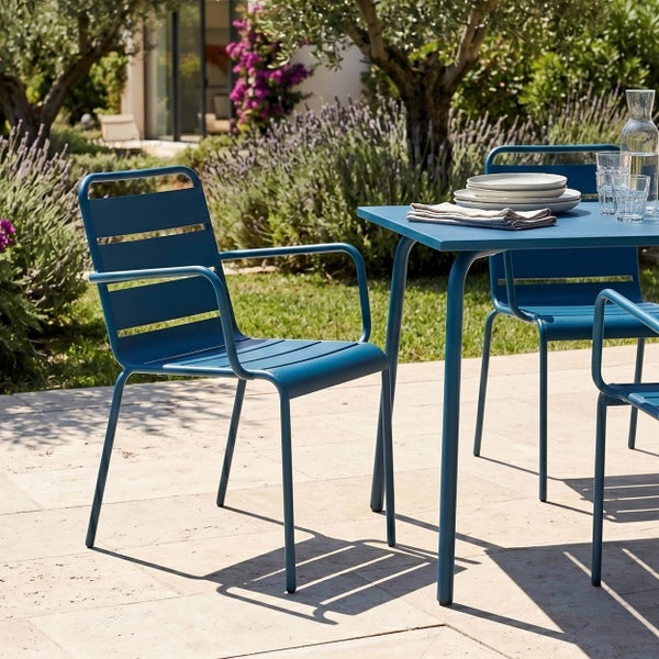 Modernes Gartenmöbel-Set aus blauem Metall auf einer Steinterrasse, bestehend aus quadratischem Tisch und Stühlen mit Armlehnen in einem Garten.