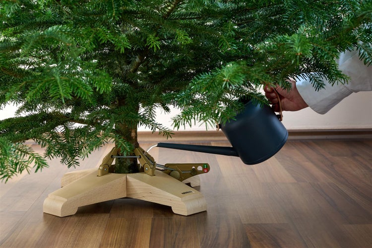 Person gießt Wasser mit einer Gießkanne in einen Christbaumständer unter einem Weihnachtsbaum.
