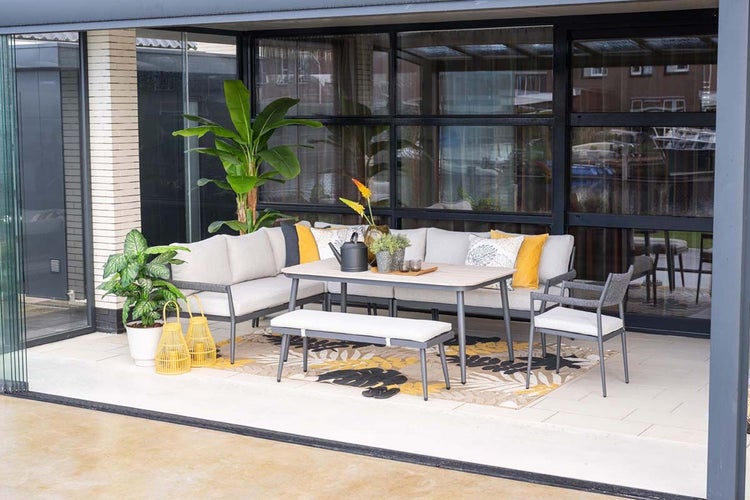 Moderne Gartenlounge bestehend aus Ecksofa, Esstisch, Sitzbank und Sessel auf einer Terrasse. Metallgestell mit hellen Polstern und Dekoration.