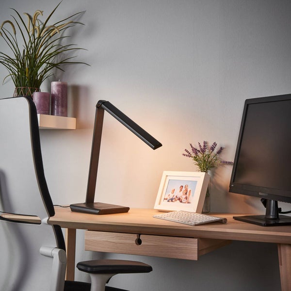 Schwarze LED-Schreibtischleuchte auf einem Holzschreibtisch in einem hellen Arbeitszimmer mit Monitor, Tastatur und Bilderrahmen.