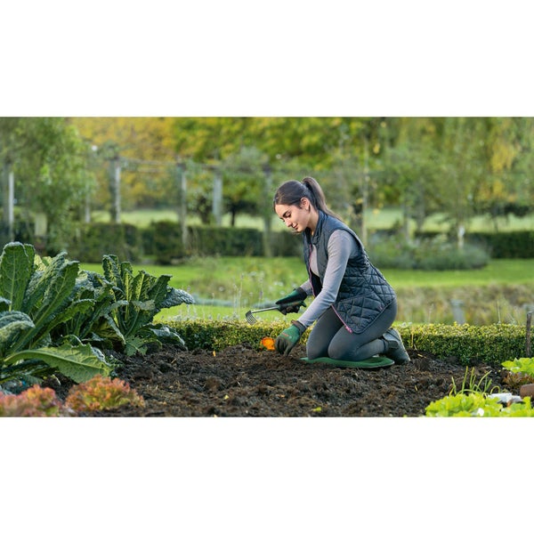 Frau kniet auf einem Kniekissen im Garten und harkt den Boden.