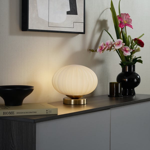 Moderne Tischleuchte mit geripptem Glasschirm und Messingfuss auf einem dunklen Sideboard neben einer Vase mit Kunstblumen und einem Buch.