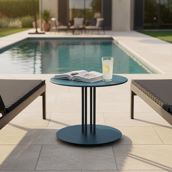 Moderner runder Beistelltisch aus Metall neben einem Pool, mit Buch und Getränk, flankiert von zwei Gartenliegen auf einer hellen Terrasse.