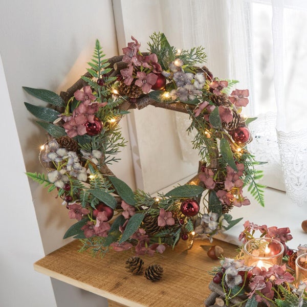 Weihnachtskranz aus Zweigen mit Kunstblumen, Beeren, Zapfen und integrierter Lichterkette als Dekoration auf einer Holzkonsole.