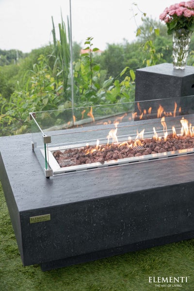 Elementi Feuertisch in rechteckiger Form mit Windschutz aus Glas, Lavasteinen und brennenden Flammen im Außenbereich.