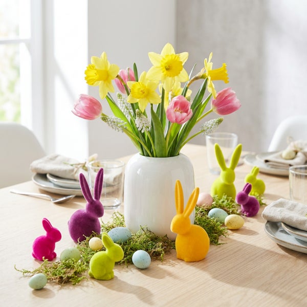 Osterdekoration auf einem Holztisch mit einer Blumenvase, bunten Osterhasenfiguren und Ostereiern in einer hellen Raumatmosphäre.