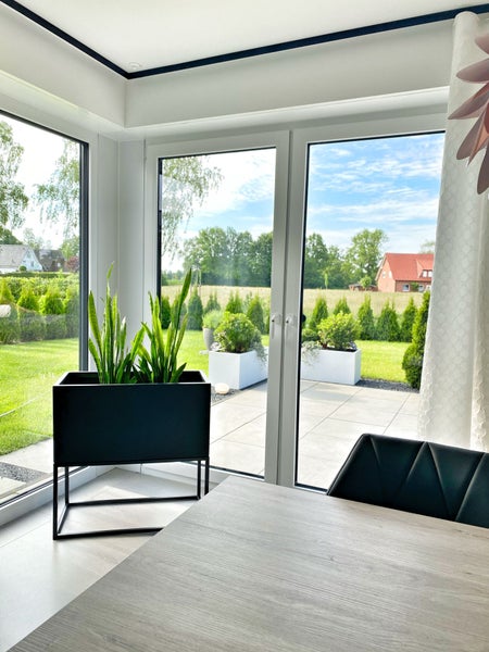 Hell gestalteter Wohnraum mit Blick durch Glastüren auf Terrasse und Garten, schwarzer erhöhter Pflanzkübel mit Bogenhanf.