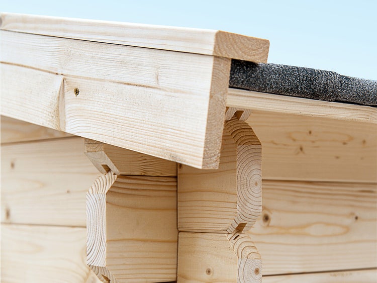 Nahaufnahme der Eckverbindung eines Gartenhauses aus hellen Blockbohlen mit Dachüberstand und dunkler Dachpappe.