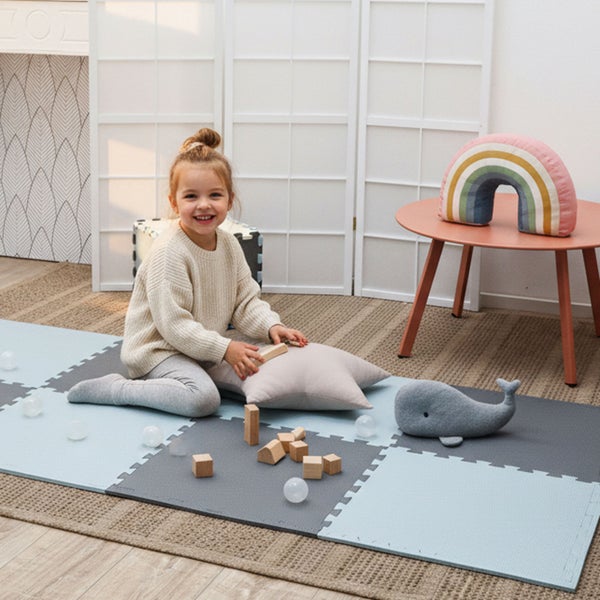 Kind spielt auf einer Puzzlematte aus Schaumstoff in einem hellen Kinderzimmer mit Holzbausteinen und Stofftieren.