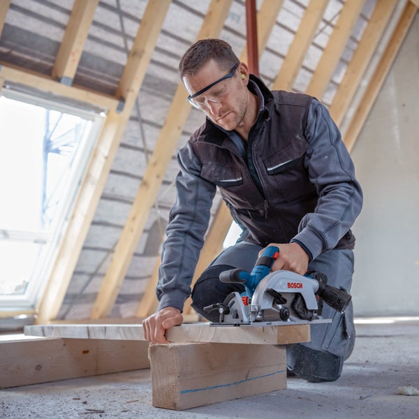 Handwerker schneidet einen Holzbalken mit einer Bosch Handkreissäge im hellen Dachgeschossrohbau unter Einhaltung von Sicherheitsvorkehrungen.