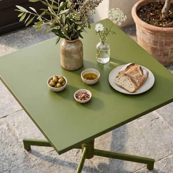 Quadratischer Gartentisch in Grün aus Metall mit Oliven, Brot und Blumenvasen auf einer Terrasse.