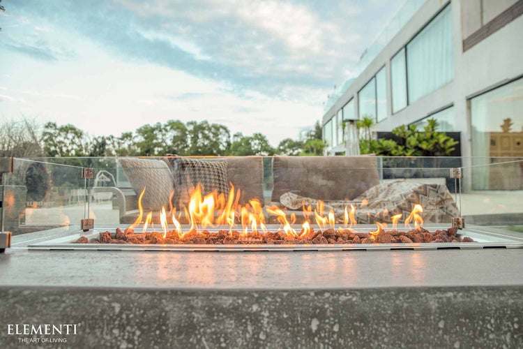 Moderne Gas-Feuerstelle mit Lavasteinen und Glas-Windschutz auf einer Terrasse mit Loungemöbeln vor einem hellen Haus, Elementi Logo.