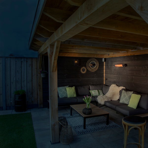 Überdachter Außenbereich mit dunkler Holzwand, Ecksofa mit Kissen, Holztisch auf einem Teppich und einem leuchtenden Infrarotstrahler.