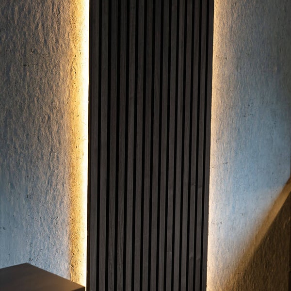 Akustikpaneel mit dunklen Holzlamellen und indirekter Beleuchtung auf einer Betonwand.