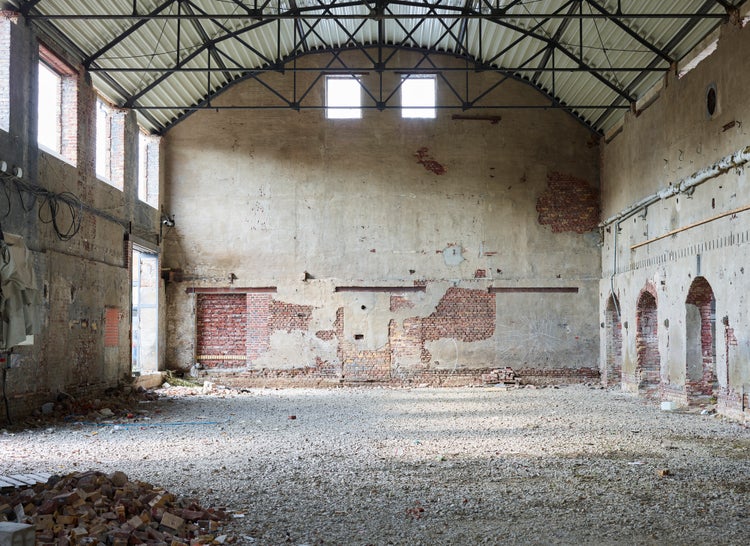 Große Industriehalle im Rohbauzustand mit freiliegenden Backsteinwänden, Metall-Dachkonstruktion und Kiesboden für Renovierungsprojekte.