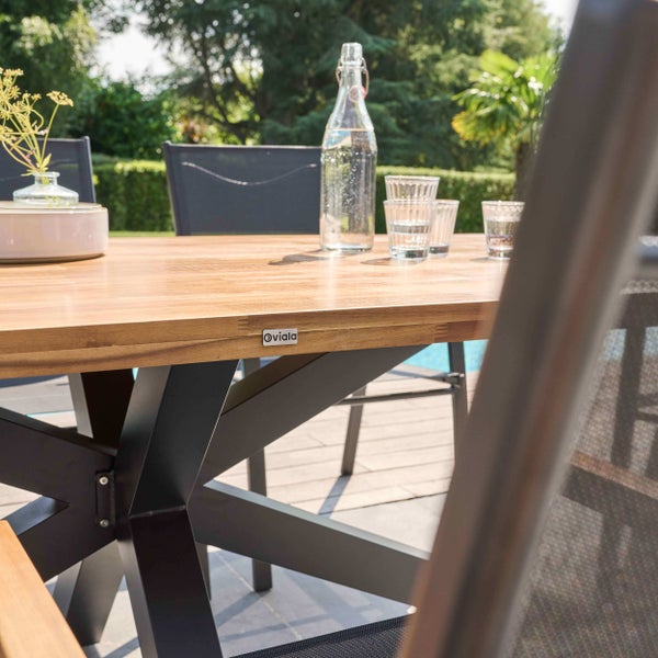 Gartentisch mit Holzplatte und schwarzem Metallgestell in X-Form, Oviala Logo, auf einer hellen Terrasse mit Pool und Sitzgelegenheiten.