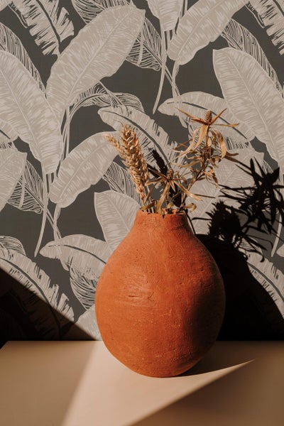 Eine runde Terrakotta Vase mit Trockenblumen steht vor einer Tapete mit einem Blattmuster.