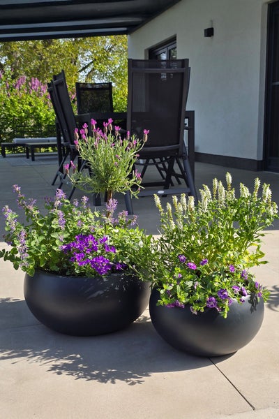 Zwei runde schwarze Pflanzkübel auf einer Terrasse, bepflanzt mit Lavendel und Sommerblumen, im Hintergrund eine Sitzgruppe mit Gartenstühlen.