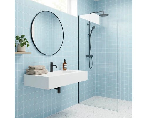 Modernes helles Badezimmer mit quadratischen Wandfliesen, weißem Waschbecken, rundem Spiegel mit schwarzem Rahmen und Dusche mit Glaswand.