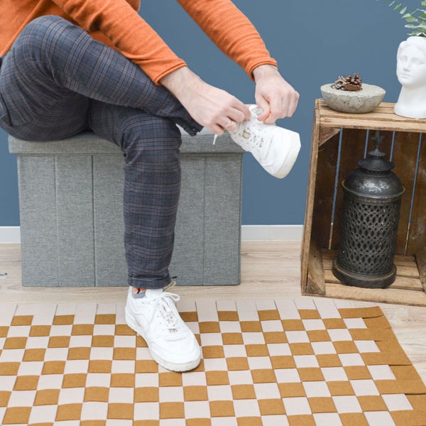 Graue gepolsterte Sitzbox mit Stauraum, auf der eine Person Schuhe bindet, davor ein gewebter Teppich mit Schachbrettmuster.