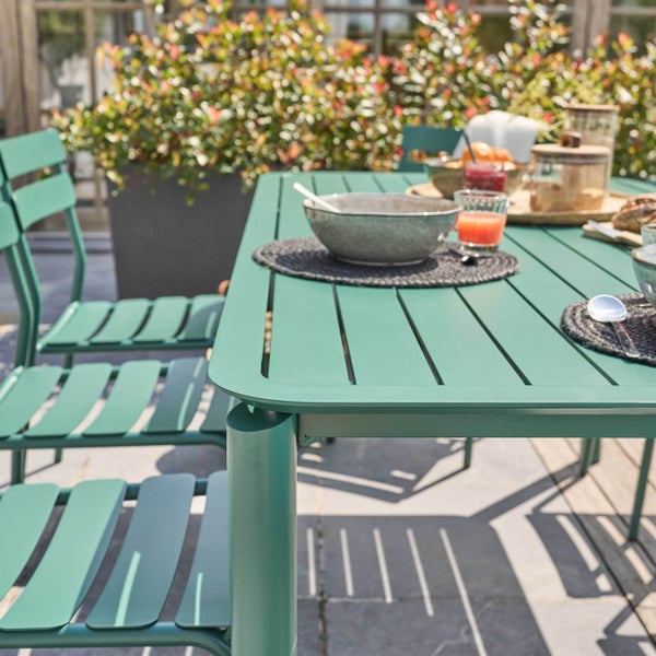 Grüner Gartentisch und Gartenstühle aus Metall in Lamellenoptik, gedeckt für ein Frühstück auf einer hellen Terrasse mit Pflanzen.