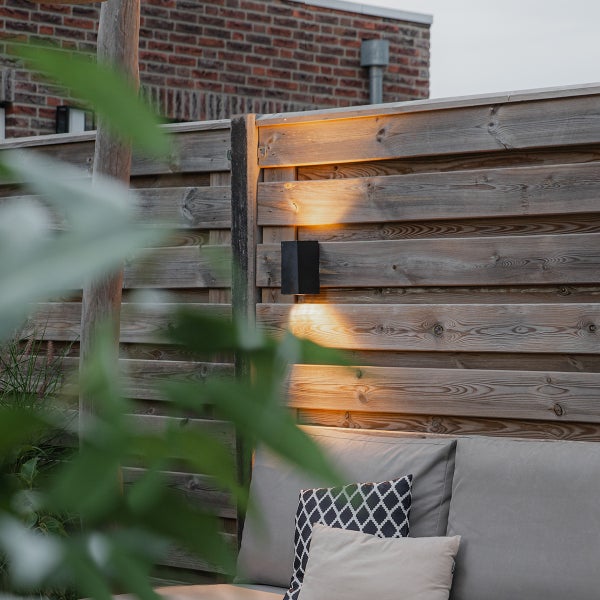 Schwarze Außenwandleuchte mit Up-and-Down-Lichteffekt an einem Holzzaun über einer Sitzecke.