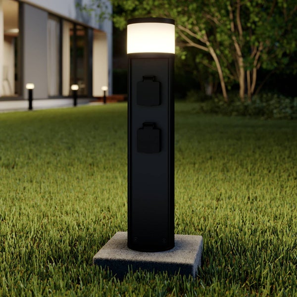 Schwarze Außenleuchte mit zwei Steckdosen und LED-Beleuchtung auf einem Rasen.