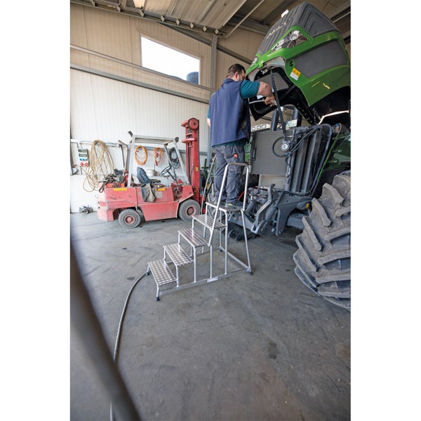 Ein Arbeiter steht auf einer Aluminium Treppenstehleiter in einer Werkstatt und repariert einen Traktor neben einem Gabelstapler.