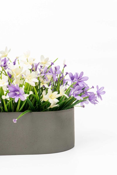 Künstliche Pflanze mit weißen und violetten Blüten in einem grauen Topf in Betonoptik.