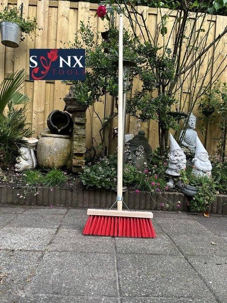 Besen mit Holzstiel und rotem Borstenkopf vor Gartenkulisse